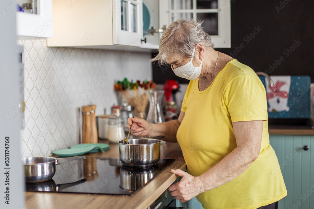 old woman in mask quarantine europe cooks in the kitchen in the apartment . Elderly at risk for coronavirus covid-19. Stay at home. live a normal life grandmother. danger of infected old woman in mask quarantine europe cooks in the kitchen in the apartment . Elderly at risk for coronavirus covid-19. Stay at home. live a normal life grandmother. danger of infected