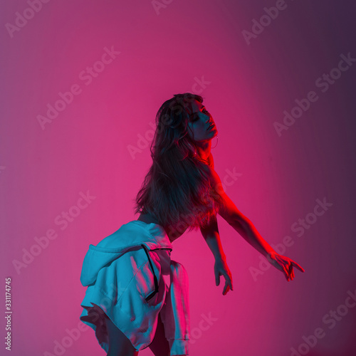 Fototapeta Athletic young woman fashion model in stylish clothes posing in a room with multi-colored glitter. Beautiful sexy girl dancer enjoys a bright pink neon in disco style background.