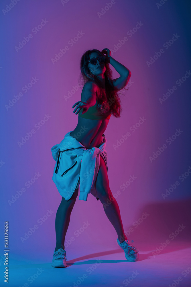 Fototapeta Young woman dancer in stylish youth clothes in sunglasses in gym shoes dancing in a room with bright neon blue-pink color.Girl hipster enjoys a dance in the studio with multi-colored ultraviolet light