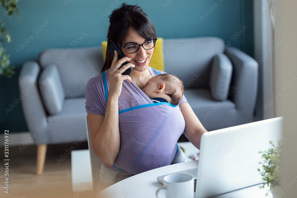Pretty young mother with her baby in sling talking on mobile phone while working with laptop at home.