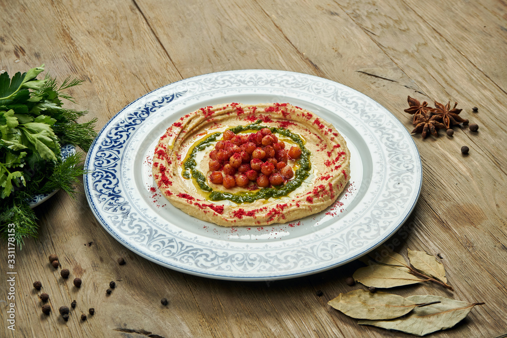 Hummus with red spices, chickpeas and herbs on a ceramic plate on a wooden background. Arabic or Israeli cuisine. Close up, horizontal. Snack Hummus with red spices, chickpeas and herbs on a ceramic plate on a wooden background. Arabic or Israeli cuisine. Close up, horizontal. Snack