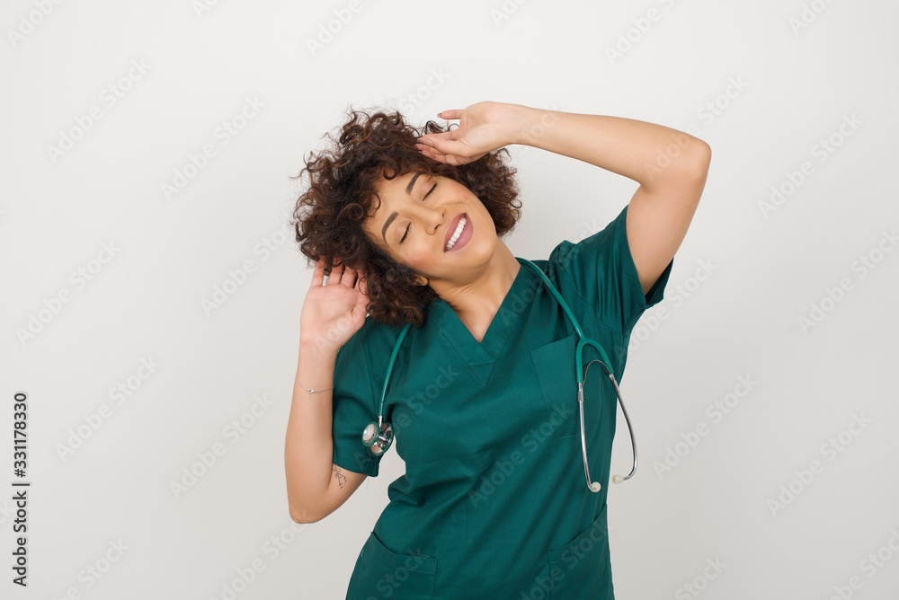 Confidence and coquettish concept. Portrait of charming young doctor girl, smiling broadly with self-assured expression while holding hands over her head. Standing against gray background. Confidence and coquettish concept. Portrait of charming young doctor girl, smiling broadly with self-assured expression while holding hands over her head. Standing against gray background.