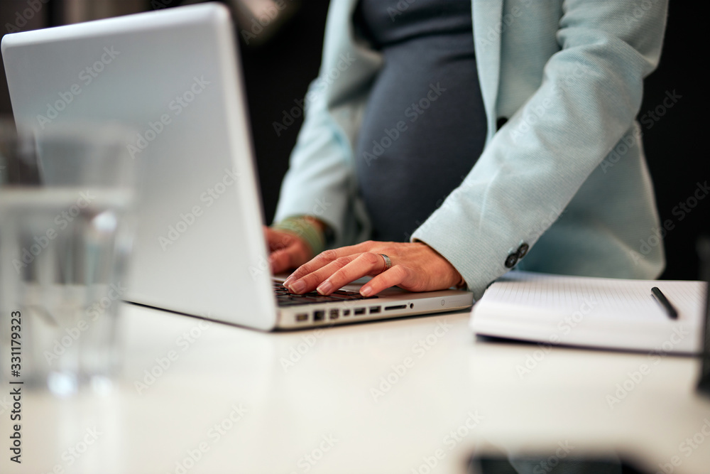 Close up of hardworking dedicated pregnant business woman standing in her office and typing report on laptop. Close up of hardworking dedicated pregnant business woman standing in her office and typing report on laptop.