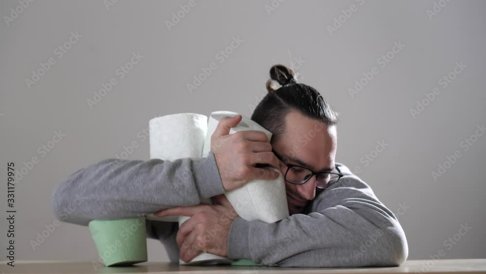 Attractive man hugs many tubes white toilet paper. Stack of toilet paper rolls against a gray background. Coronavirus or COVID-19. Pandemic worldwide, panic. Attractive man hugs many tubes white toilet paper. Stack of toilet paper rolls against a gray background. Coronavirus or COVID-19. Pandemic worldwide, panic.