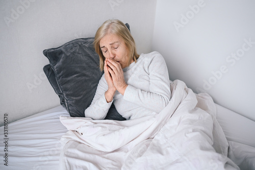 Senior woman sneezes sick of coronavirus viral infection spreading corona virus covering mouth and nose. Painful cough ill elderly patient lying in bed at home quarantine. First symptoms 