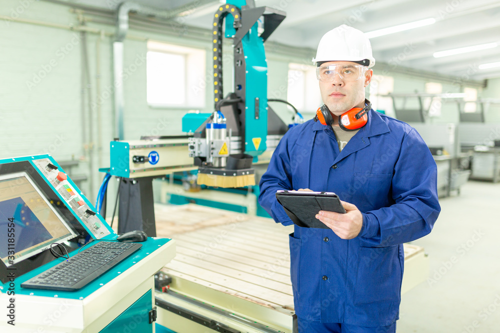 CNC machine operator in the Hard white Hat Walks Through Light Modern Factory While Holding tablet. Successful, Handsome Man in Modern Industrial Environment. CNC machine operator in the Hard white Hat Walks Through Light Modern Factory While Holding tablet. Successful, Handsome Man in Modern Industrial Environment.