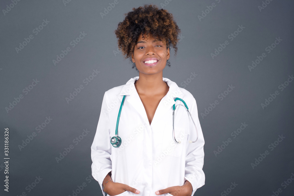 Confidence and business concept. Portrait of charming successful young doctor girl, smiling broadly with self-assured expression while holding hands in pockets standing against gray wall. Confidence and business concept. Portrait of charming successful young doctor girl, smiling broadly with self-assured expression while holding hands in pockets standing against gray wall.