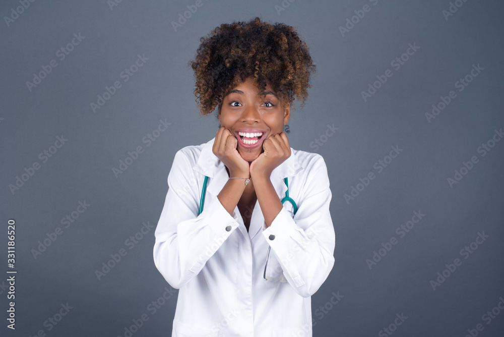 Studio shot of pleased lovely doctor woman keeps hands together under chin, being in high spirit, likes friends idea for next day, isolated on gray wall. Positive emotions. Studio shot of pleased lovely doctor woman keeps hands together under chin, being in high spirit, likes friends idea for next day, isolated on gray wall. Positive emotions.