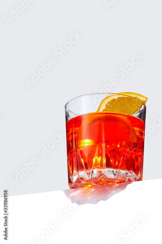 Negroni cocktail is contained in a low crystal glass with orange slices and ice cubes and isolated on a table edge. The showy illustrative picture is made on the gray backdrop.