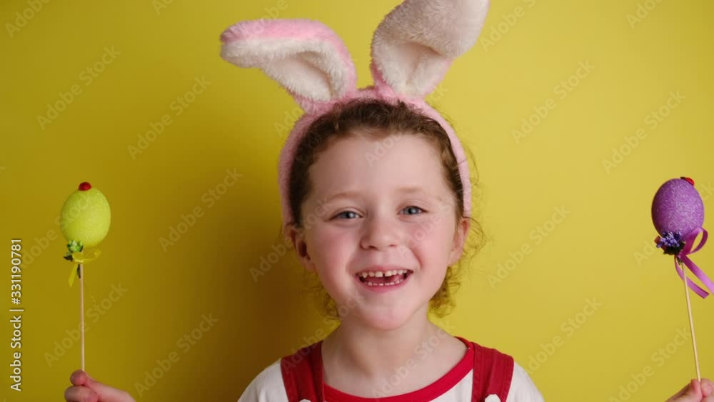 Glad cheerful little girl covers eyes with two Easter eggs, wears bunny ears, dressed in white t-shirt, models over yellow wall with free space for promotion. Spring holiday and Easter concept. Glad cheerful little girl covers eyes with two Easter eggs, wears bunny ears, dressed in white t-shirt, models over yellow wall with free space for promotion. Spring holiday and Easter concept.