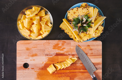 zenithal view of pineapple cut into pieces on wooden board with knife, a bowl with sliced pineapple and a plate with the remains