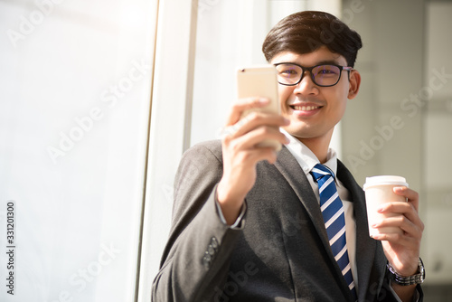 A asian handsome young businessman holding hot coffee cup while using his phone and standing on the escalator. Business concept.