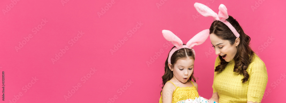 panoramic shot of surprised mother and daughter in bunny ears looking at easter eggs isolated on pink panoramic shot of surprised mother and daughter in bunny ears looking at easter eggs isolated on pink