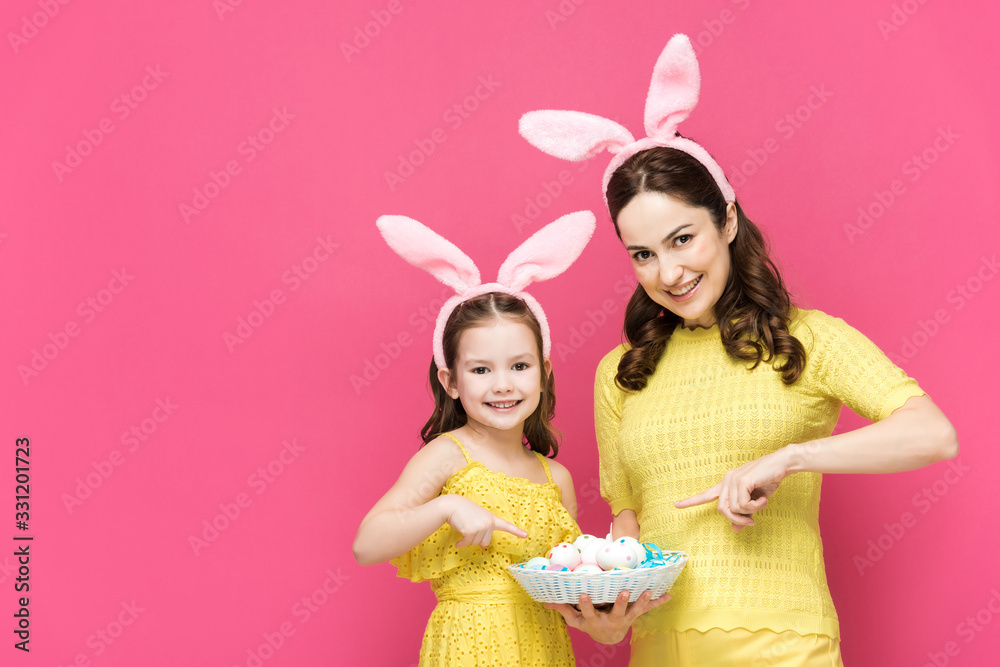 happy mother and daughter in bunny ears pointing with fingers at easter eggs isolated on pink