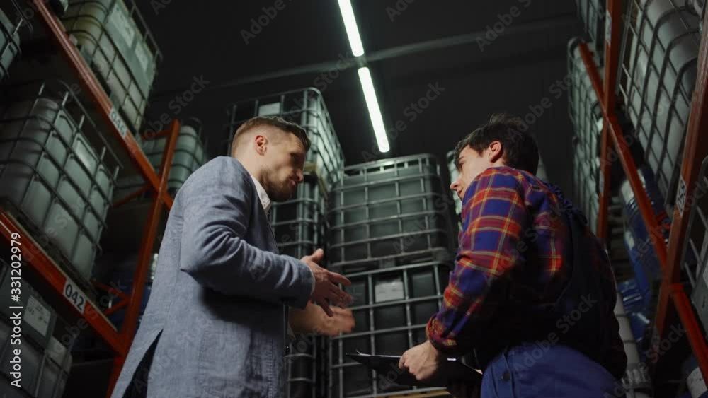 From below tracking shot of inspector or manager explaining details to young material handler in industrial storage From below tracking shot of inspector or manager explaining details to young material handler in industrial storage