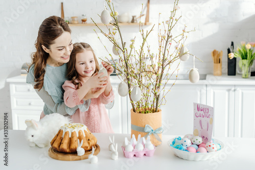 Photography Happy kid touching decorative easter egg on willow branch near mother