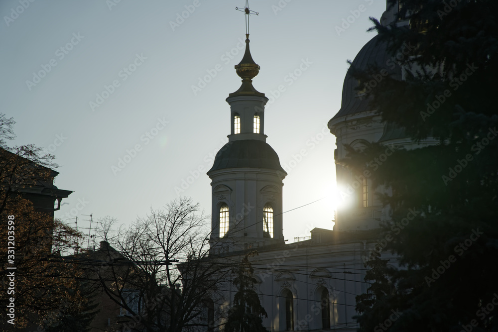 Part of ortodox church in front view with sun behind the clounds. The sun rays breaks through the dome of the church. Cross of church against blue sky.
