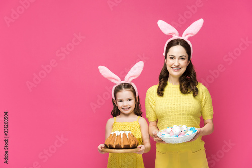 happy mother in bunny ears holding painted chicken eggs near daughter with easter cake isolated on pink
