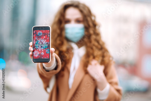 Girl in protective sterile medical mask on her face holds out the phone in quarantine city. Coronavirus cells on phone display. The concept of preventing the spread of the epidemic. 