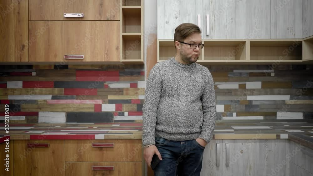 A man with a beard and glasses, dressed in jeans and a sweater stands in the kitchen and looks left and right. He chooses which kitchen to buy. A man with a beard and glasses, dressed in jeans and a sweater stands in the kitchen and looks left and right. He chooses which kitchen to buy.