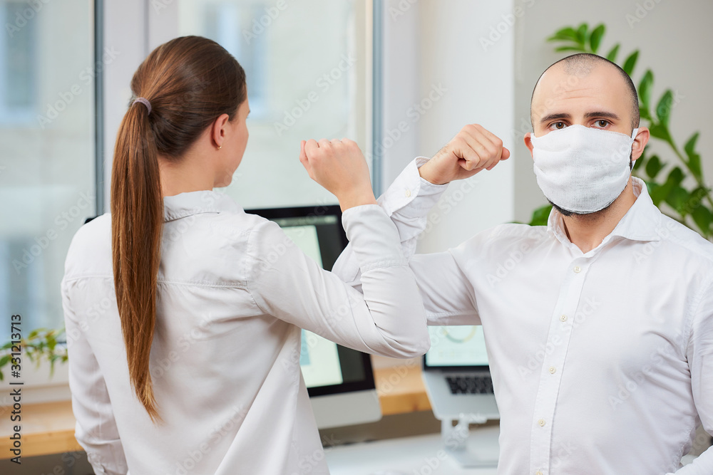Elbow greeting to avoid the spread of coronavirus (COVID-19). Man with a medical face mask and woman meet in an office with bare hands. Instead of greeting with a hug or handshake, they bump elbows Elbow greeting to avoid the spread of coronavirus (COVID-19). Man with a medical face mask and woman meet in an office with bare hands. Instead of greeting with a hug or handshake, they bump elbows
