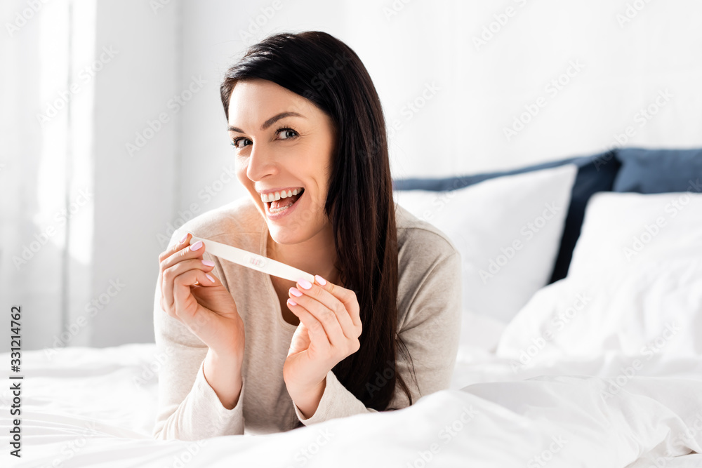 cheerful woman holding pregnancy test and lying on bed cheerful woman holding pregnancy test and lying on bed