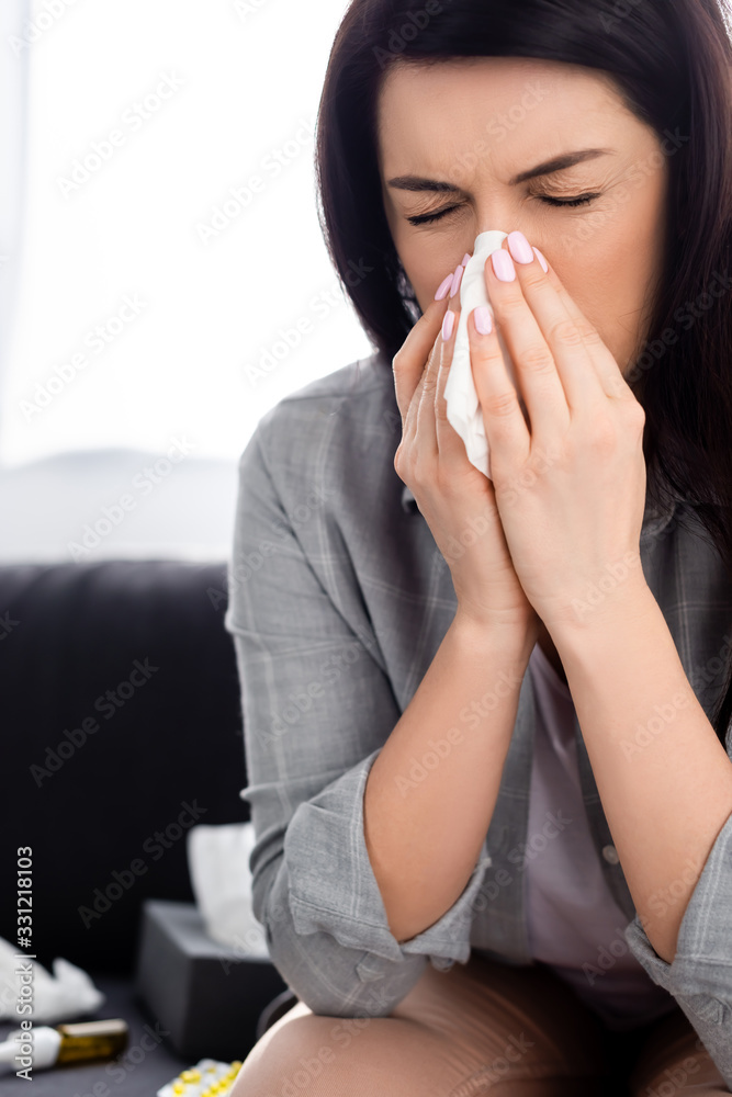allergic woman with closed eyes sneezing in napkin while sitting on sofa allergic woman with closed eyes sneezing in napkin while sitting on sofa