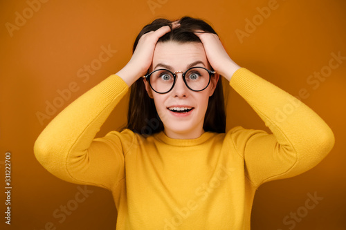 Indoor shot of surprised woman in eyeglasses expresses shock, keeps hands near head, jaw dropped and looks at camera, dressed in sweater, standing isolated on orange background. Positive human emotion