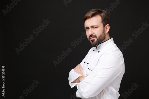 Side view of displeased young bearded male chef cook or baker man in white uniform shirt isolated on black background. Cooking food concept. Mock up copy space. Holding hands crossed, looking camera.