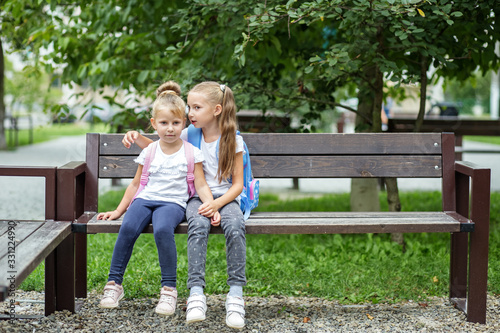 Two small children tell secrets on a bench. Girls with backpacks. The concept is back to school, family, friendship and childhood. © Olha Tsiplyar