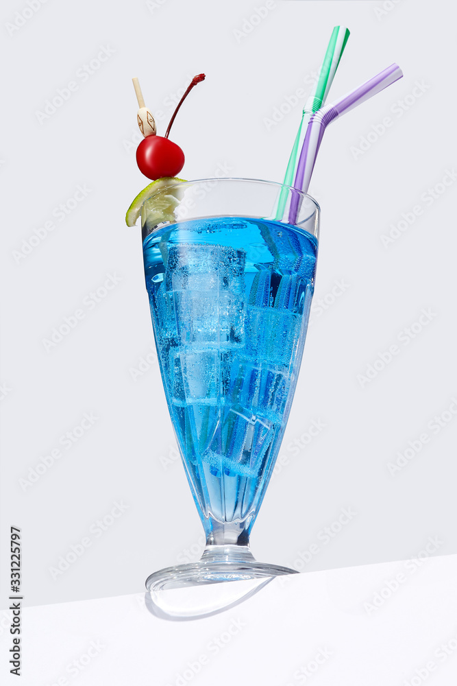 Blue Lagoon cocktail with ice cubes is contained in a milkshake glass with two straws, a lime slice and a cherry on the stick. The showy illustrative picture is made on the gray backdrop. Blue Lagoon cocktail with ice cubes is contained in a milkshake glass with two straws, a lime slice and a cherry on the stick. The showy illustrative picture is made on the gray backdrop.