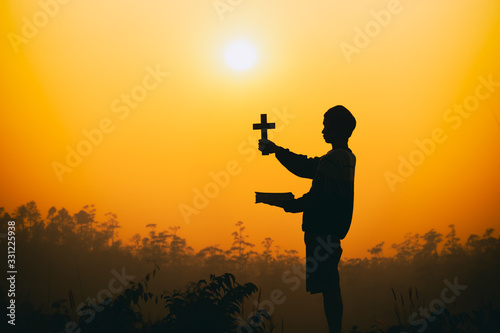 Young man holding bible ang christian cross on the mountain at sunset background. christian silhouette concept.