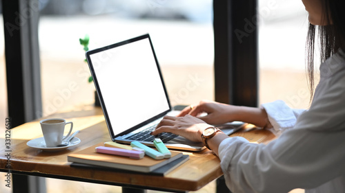 Cropped image of confident woman working with white blank screen computer laptop while sitting at the wooden working desk over modern office glass wall as background.