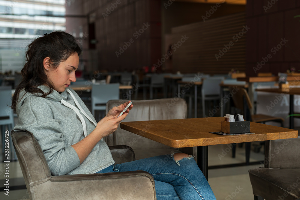 A girl with a serious expression sits alone in a cafe with a phone in her hands. Close-up. Horizontal side view.