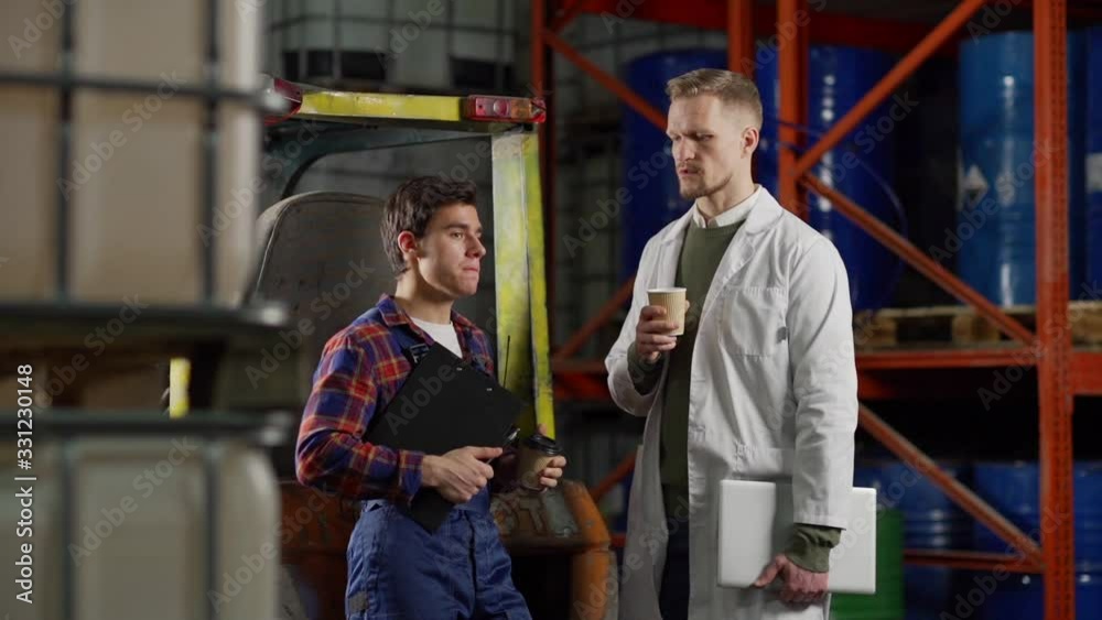 Tracking slow motion shot of middle aged engineer in white coat with laptop and young material handler with clipboard and walkie-talkie talking and drinking coffee by forklift in industrial storage