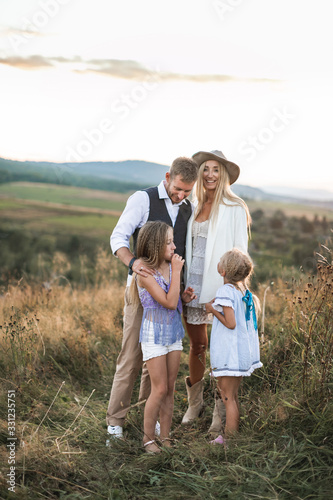 Happy smiling young family, father, mother and two little daughters having fun outdoors, standing together in a wild field. Mom and Dad talking with their kids, laughing, enjoying time together