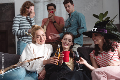 handsome men standing near cheerful girls drinking beer, wine and soda while sitting on sofa