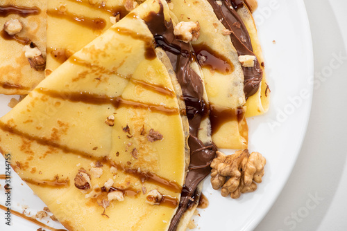 close up view of tasty crepes with chocolate spread and walnuts on plate on white background