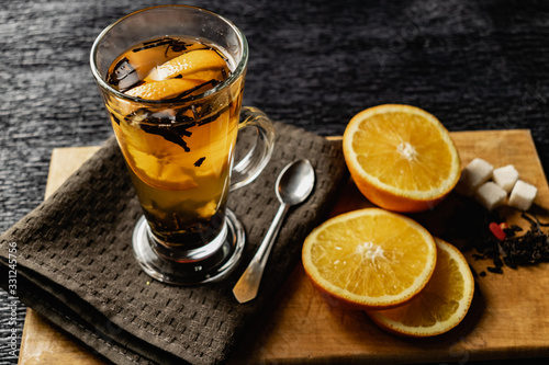 Citrus drink, tea with petals in a transparent glass mug with slices of sweet orange and refined sugar. Slicing fruit on a wooden Board with a brown napkin in a cozy dark background in a low key