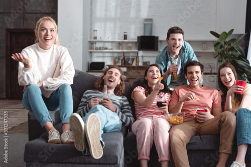 cheerful friends smiling while sitting on sofa at home and watching tv