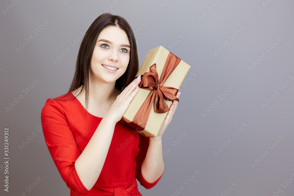 Lovely girl 15-20 years old in a red dress with a gift. A brunette girl in a bright red dress on gray background holds a gift with a ribbon in her hands. Lovely girl 15-20 years old in a red dress with a gift. A brunette girl in a bright red dress on gray background holds a gift with a ribbon in her hands.