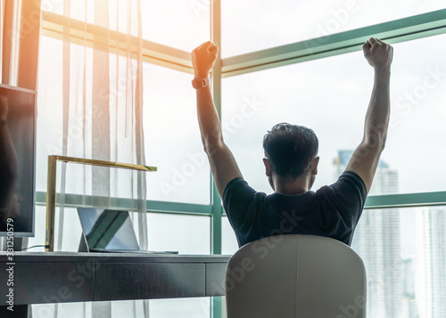 Business achievement concept with happy businessman relaxing in office or hotel room, resting and raising fists with ambition looking forward to city building urban scene through glass window