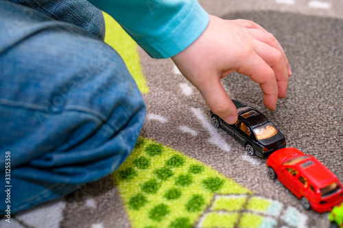 Close-up of five year old boy playing and lining up toy cars on a playing mat with roads. The cars have vivid colors and the boy is dressed in blue jeans and a blue shirt. He is picking up a black toy