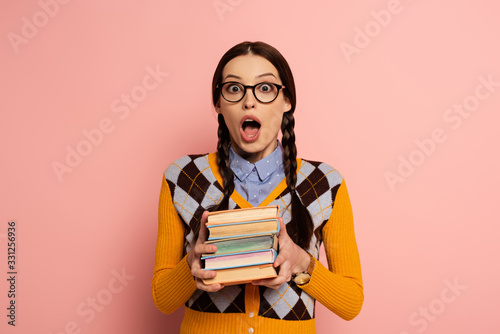 shocked female nerd with open mouth holding books on pink