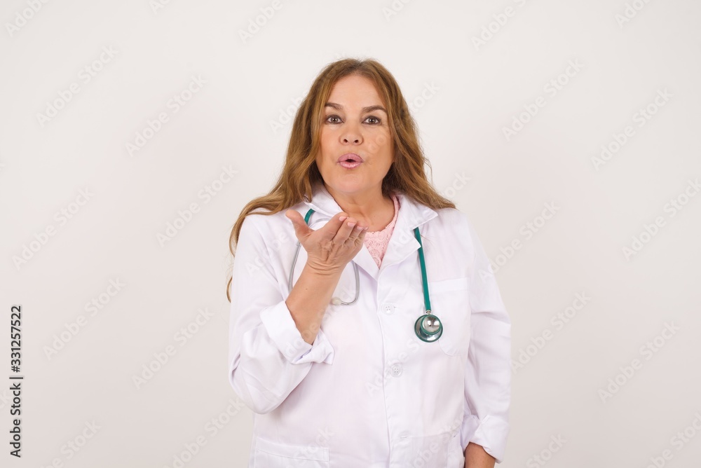 Cute appealing doctor female wearing medical uniform blows kiss at camera, demonstrates love to boyfriend or says goodbye on distance, isolated over studio background. Cute appealing doctor female wearing medical uniform blows kiss at camera, demonstrates love to boyfriend or says goodbye on distance, isolated over studio background.
