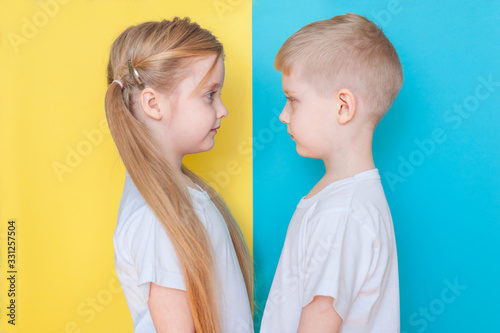 Blond twins boy and girl in white T-shirts stand and look at each other on blue and yellow backgrounds.