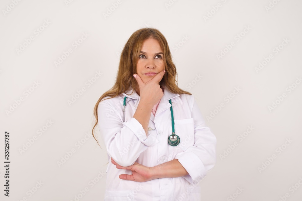 Isolated portrait of stylish young Caucasian doctor woman with hand under chin and looking sideways with doubtful and skeptical expression, worry and doubt. Standing indoors over background. Isolated portrait of stylish young Caucasian doctor woman with hand under chin and looking sideways with doubtful and skeptical expression, worry and doubt. Standing indoors over background.