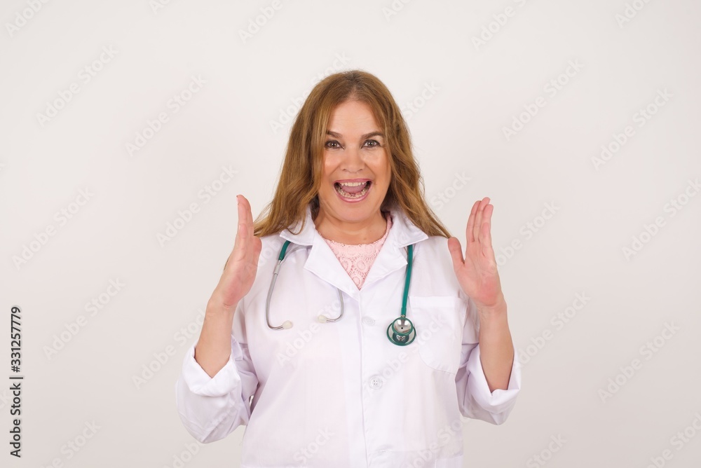 Portrait of beautiful doctor female looks with excitement at camera, keeps hands raised over head, notices something unexpected, isolated over gray wall. Lovely woman reacts on sudden news. Portrait of beautiful doctor female looks with excitement at camera, keeps hands raised over head, notices something unexpected, isolated over gray wall. Lovely woman reacts on sudden news.