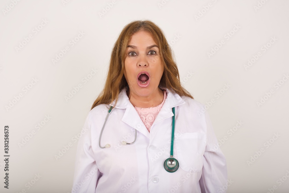 Expressive facial expressions. Shocked stupefied doctor woman wearing medical uniform, feels stunned from what she sees aside on blank gray Wall