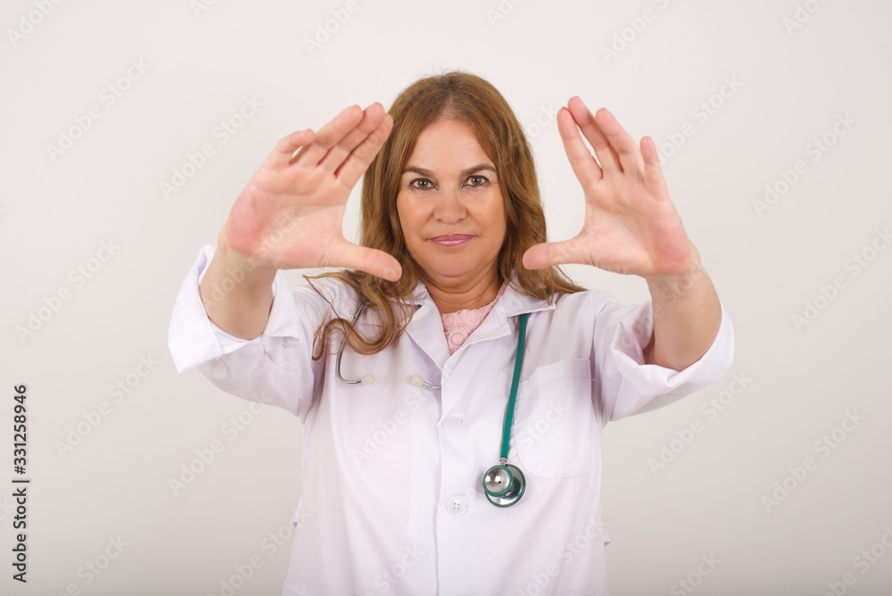 Gesturing finger frame. Portrait of smiling beautiful young doctor woman looking at camera and gesturing finger frame while standing indoors. Gesturing finger frame. Portrait of smiling beautiful young doctor woman looking at camera and gesturing finger frame while standing indoors.